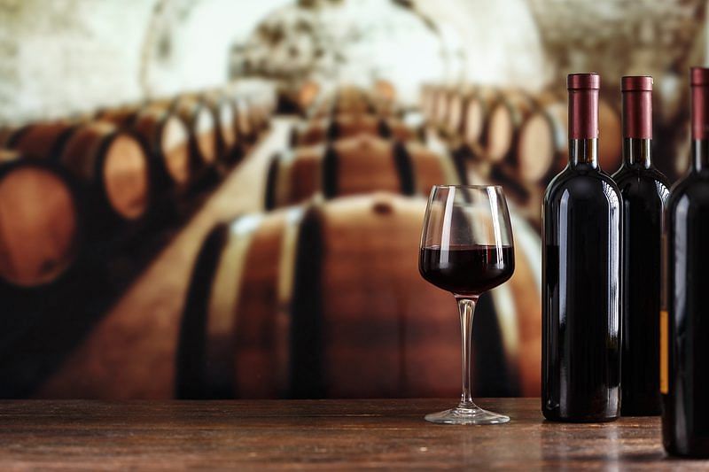 shot of a glass of wine with wine bottles beside it and a blurred background of wine barrels