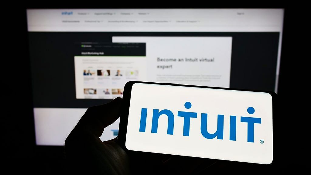 A logo of Intuit on a screen of a horizontally flipped smartphone; at the back is a computer screen showing Intuit’s website homepage.