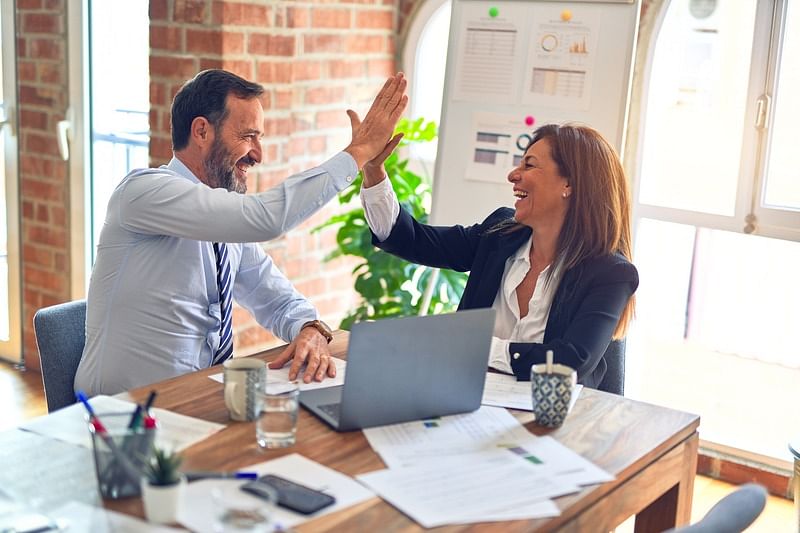 Two business people sitting opposite each other does high five in front of a laptop on a wooden table cluttered with documents.